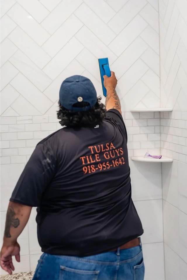 Craftsman applying grout to a white herringbone tile shower wall beside a corner shelf